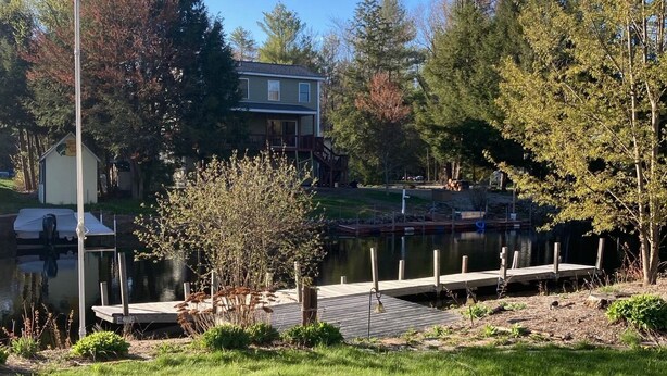A grassy area with a dock extending into a body of water, surrounded by trees and a building with a balcony. - Lakefront Home for Rent (Moultonborough)