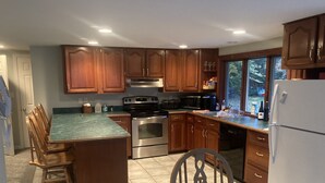 A kitchen with wooden cabinets, a stainless steel oven, a refrigerator, and a dining area with chairs and a table. - Lakefront Home for Rent (Moultonborough)