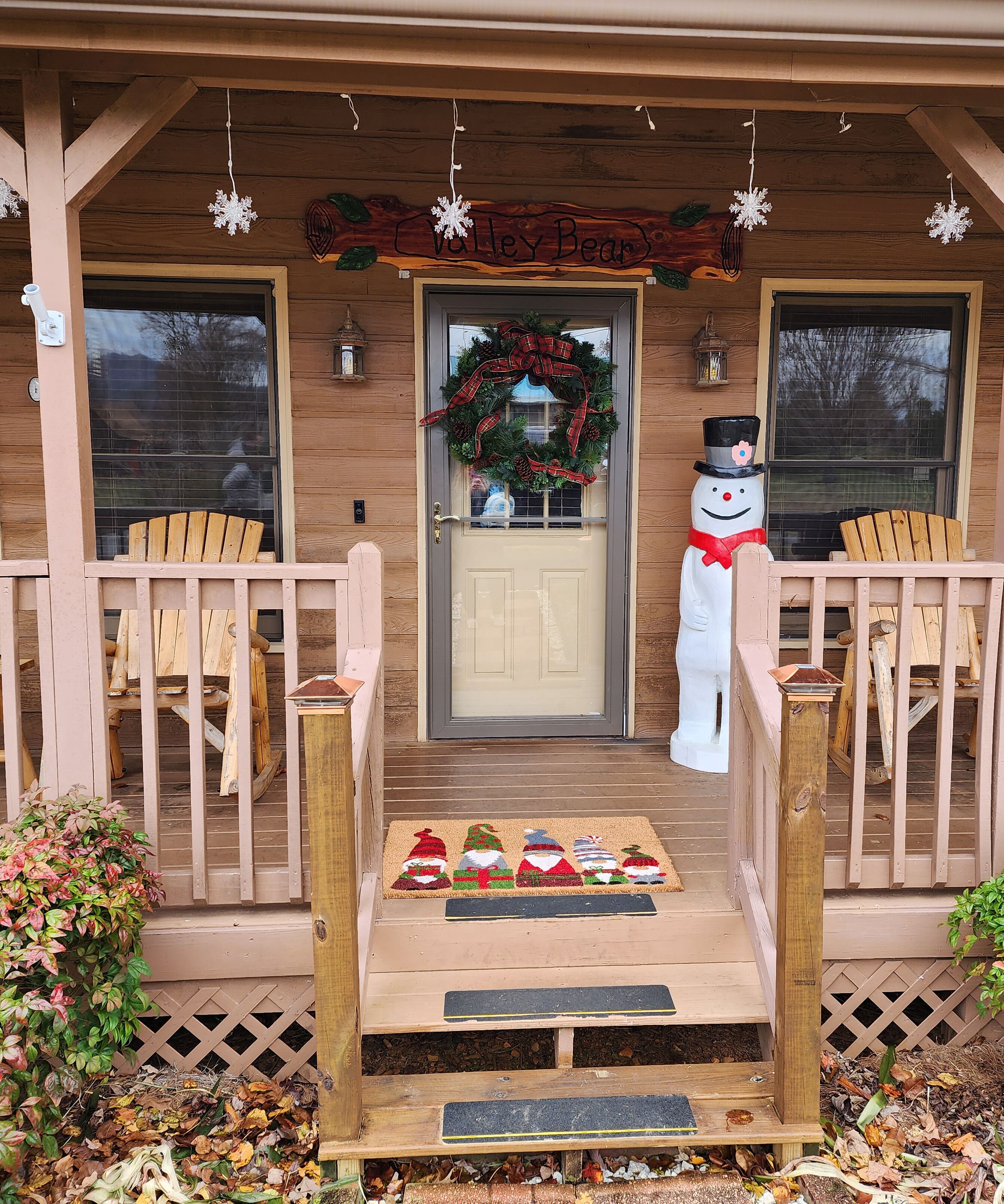 A wooden porch with a welcome mat featuring Santa Claus and snowmen, a snowman decoration, and a wreath on the door.