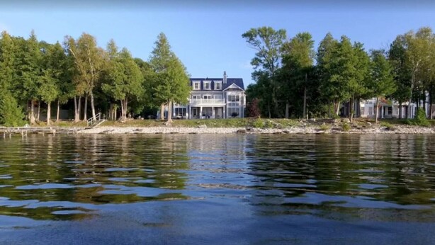 Une propriété située au bord d’un lac, avec une grande maison dotée de nombreuses fenêtres et d’un balcon, entourée d’arbres et bordée par une étendue d’eau paisible.