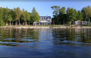 Propiedad frente a un lago con una casa grande de múltiples ventanales y balcón, rodeada de árboles y una masa de agua tranquila.