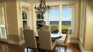 Un coin repas avec une table ronde et six chaises, de grandes fenêtres donnant sur la mer et un lustre.