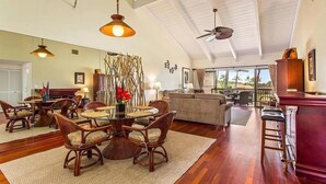 A spacious living room with a high ceiling, a dining area with wicker furniture, and a view of the outdoors through large windows. - Spacious Executive Retreat in Paradise + 3 mins from Turtle Beach! (Kailua-Kona)