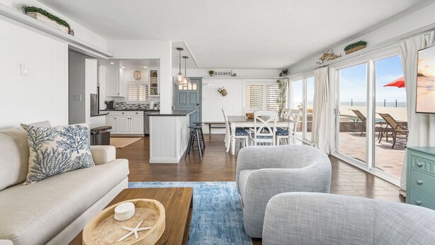 Una sala de estar moderna con un sofá, una mesa de centro y un comedor con una mesa y sillas. Se ve una cocina al fondo y una vista de la playa a través de amplios ventanales.