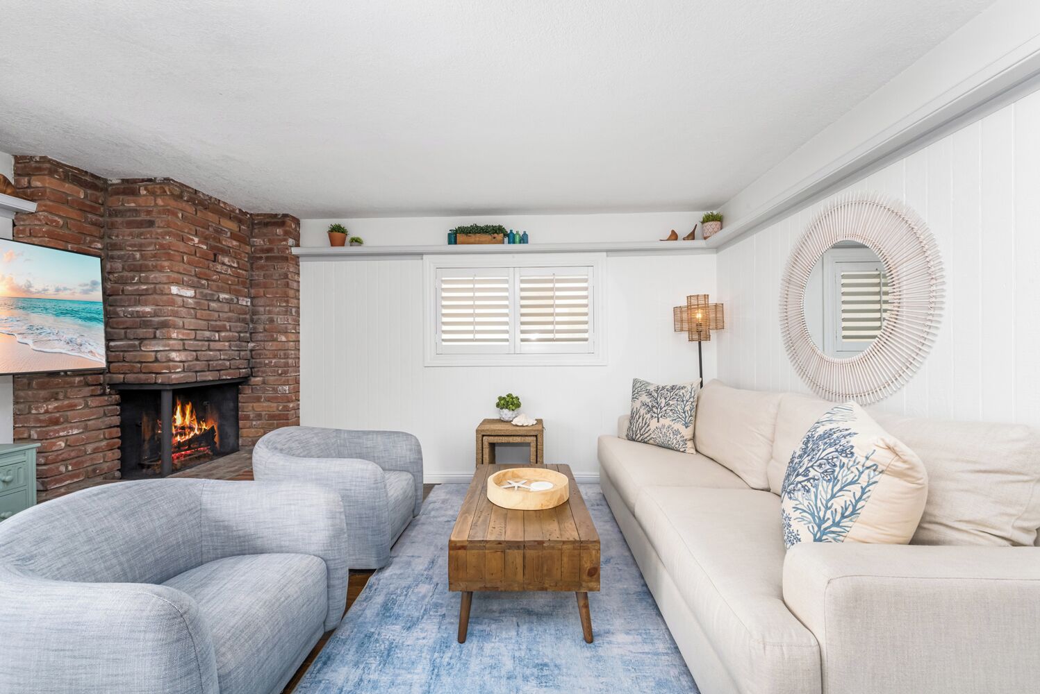 A cozy living room with a brick fireplace, two grey sofas, a wooden coffee table, and a white wall with a mirror and window blinds.