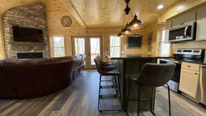 Un intérieur de chalet chaleureux avec une cheminée en pierre, un plafond en bois, un coin bar avec des tabourets, une cuisine équipée d’appareils modernes et une grande fenêtre.