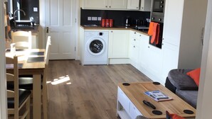 A compact living space with a kitchen, a washing machine, a dining area, and a seating area with a sofa and a coffee table. - Studio (Lochinver)