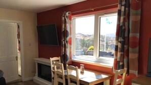 A dining area with a table and chairs, a fireplace, a TV mounted on the wall, and a window with curtains. - Studio (Lochinver)