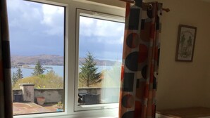 A bedroom with a bed, a chair, a window with a view of a lake and hills, and a curtain. - Studio (Lochinver)