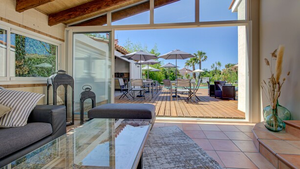 A spacious living area with a glass coffee table, a sofa, and a view of an outdoor patio with umbrellas and outdoor furniture. - Domaine Le Courti (Le Barcares)
