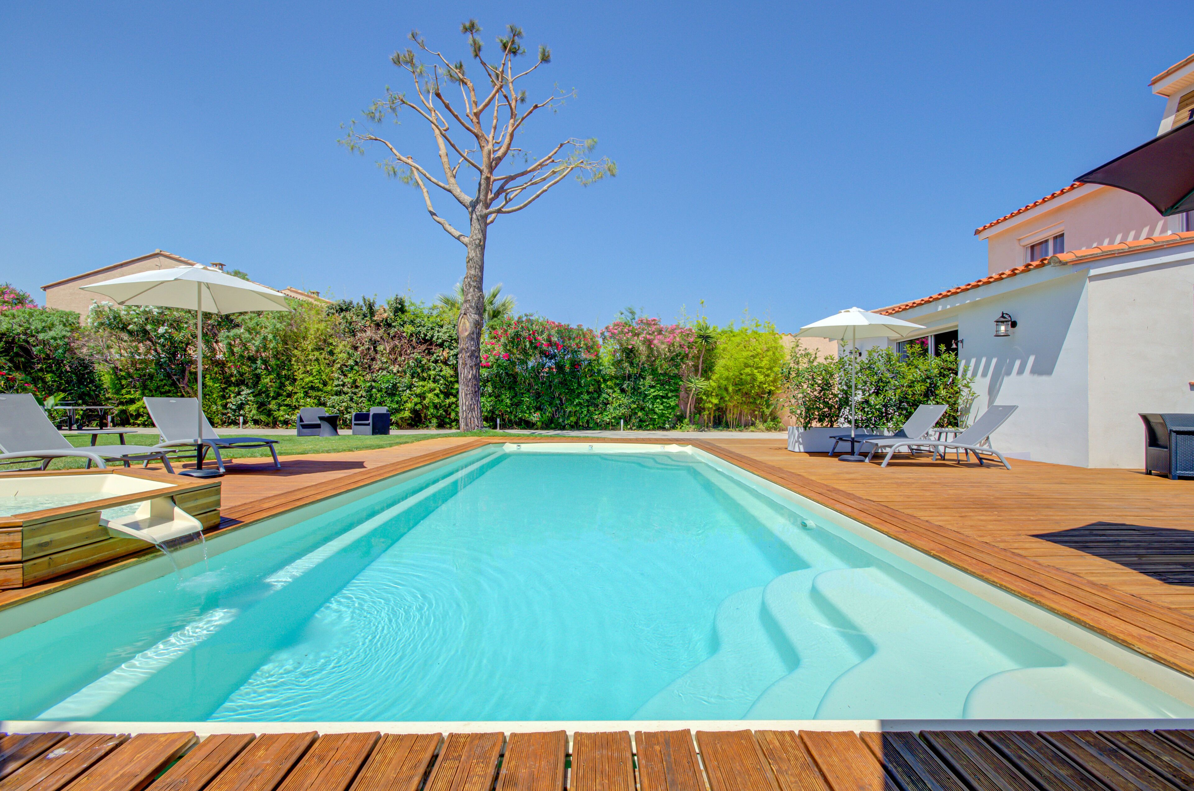 A rectangular swimming pool with wooden deck flooring, surrounded by lounge chairs and umbrellas.