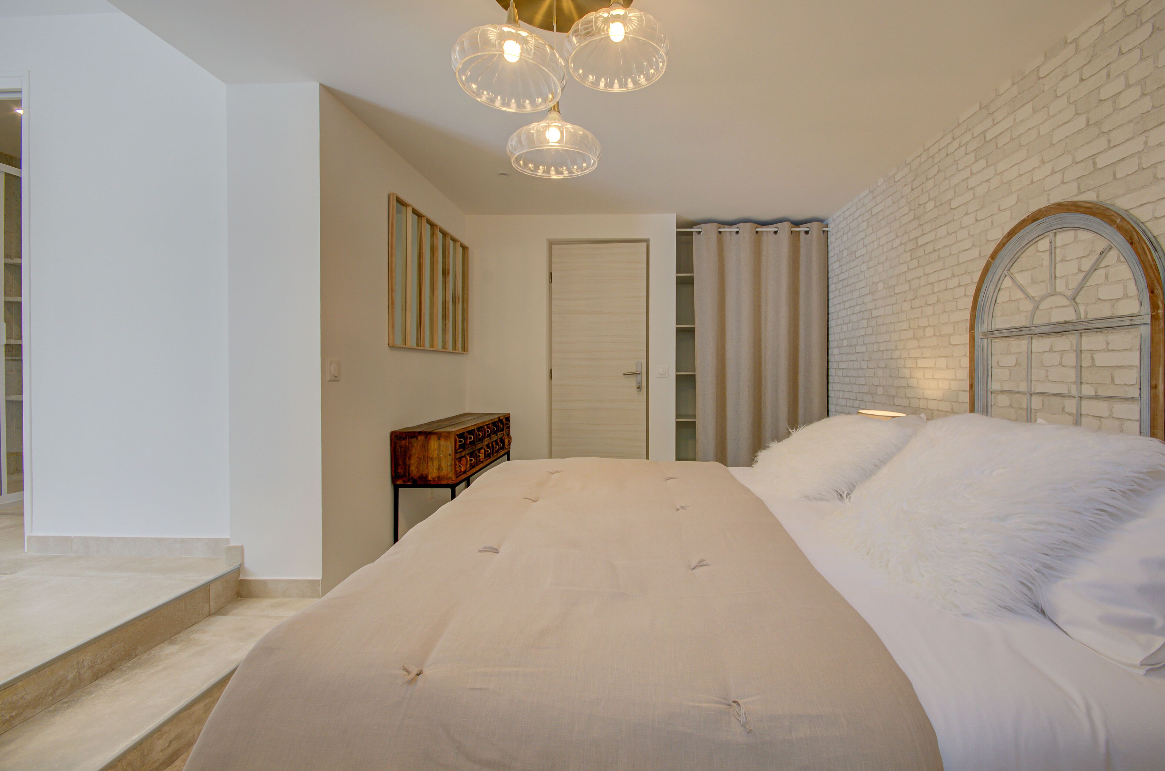 A bedroom with a large bed, a wooden nightstand, a white door, and a brick wall with a window.