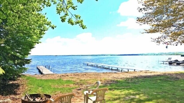 A lakeside view with a dock extending into the water, a grassy area with a fire pit and plastic chairs, and a clear sky. - Brothers lake drive chalet (Shawano)