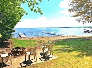 Blick auf einen See mit einem Steg, der ins Wasser führt, einer Wiese mit Feuerstelle und Plastikstühlen, und einem klaren Himmel.