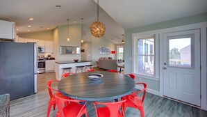 A modern kitchen with a round table and red chairs, a refrigerator, and a dining area with a view of the outside. - Double Dog Dare – Sound-View OBX Retreat in Black Dog Harbor, Salvo (SALVO)