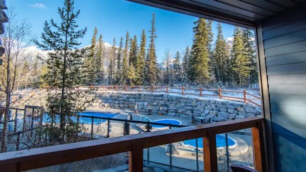 A view from a room with a wooden railing, overlooking a pool area with a stone wall, surrounded by trees and a mountain in the background. - Lux Blackstone Mountain Suite with Amazing Views (Canmore)