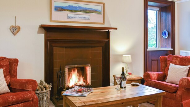 Un salon chaleureux avec une cheminée, deux canapés rouges à motifs, une table basse en bois et, au-dessus du manteau de cheminée, un tableau représentant un paysage encadré.