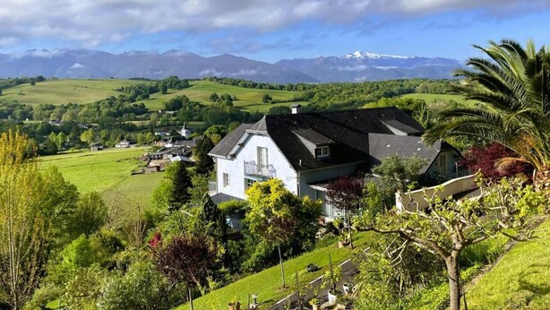 Uma casa com telhado inclinado, cercada por vegetação exuberante e vista para montanhas ao longe.
