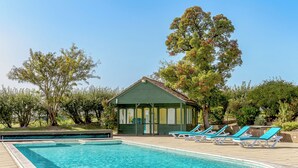 Piscina com espreguiçadeiras, um pequeno edifício e árvores.