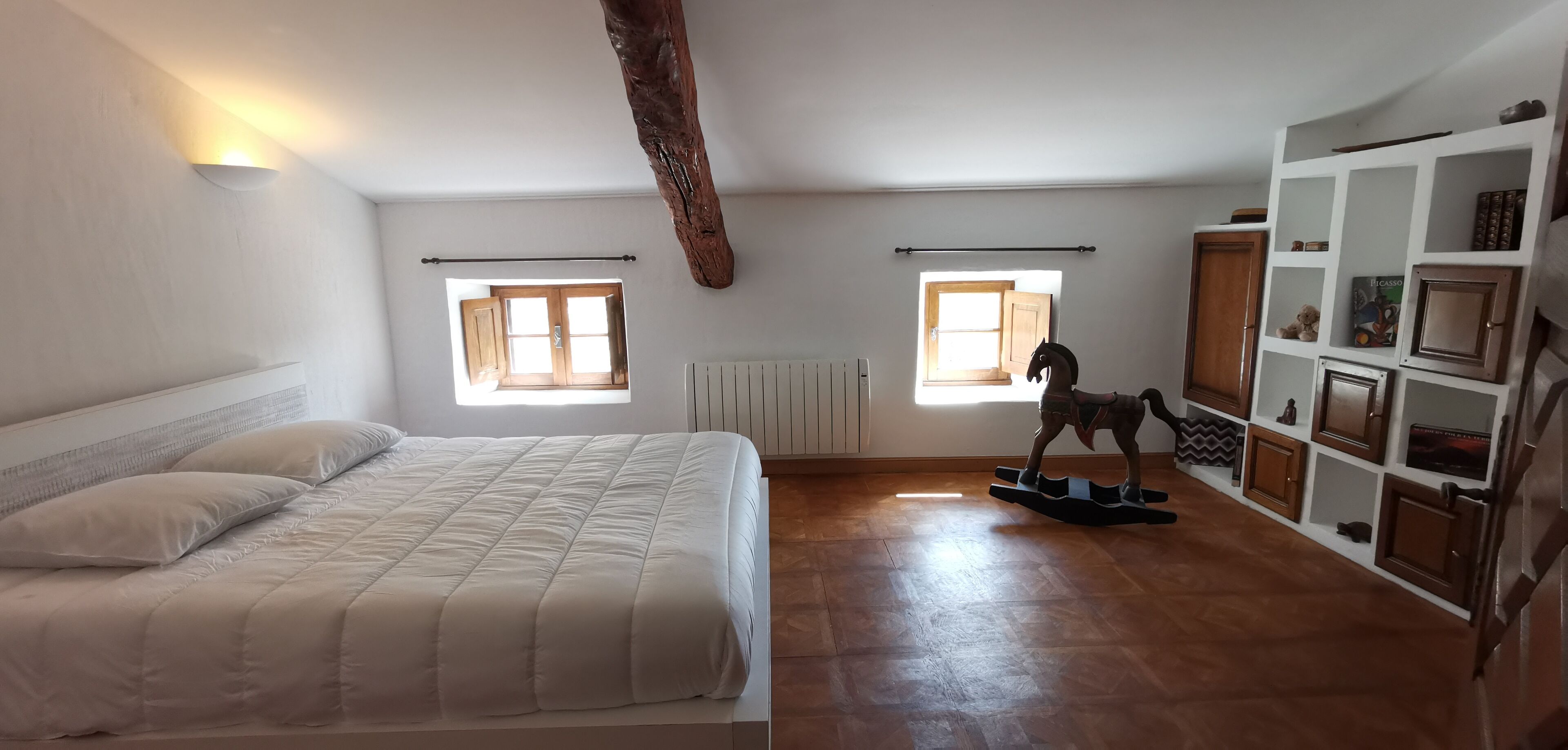 Ein Schlafzimmer mit einem Bett, zwei Fenstern, einem Heizkörper, einem Holzbalken und einem Regal mit verschiedenen Gegenständen.