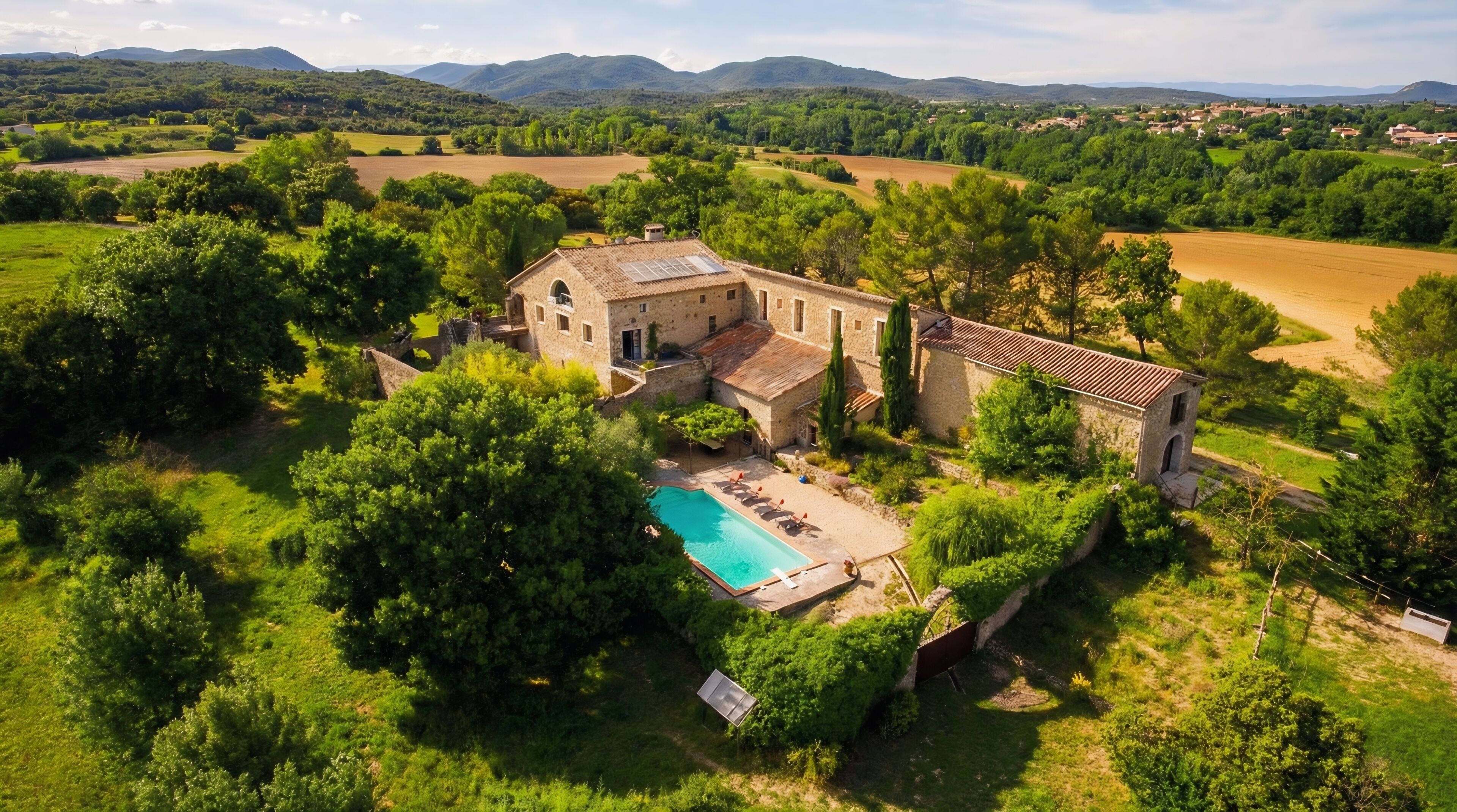Un grand bâtiment en pierre doté d’une piscine et d’un espace extérieur aménagé, entouré d’une végétation luxuriante et de champs.