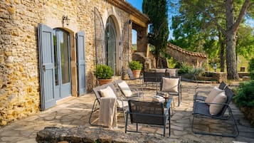Un bâtiment en pierre avec des volets bleus, des sièges d’extérieur avec des coussins et des plantes en pot.