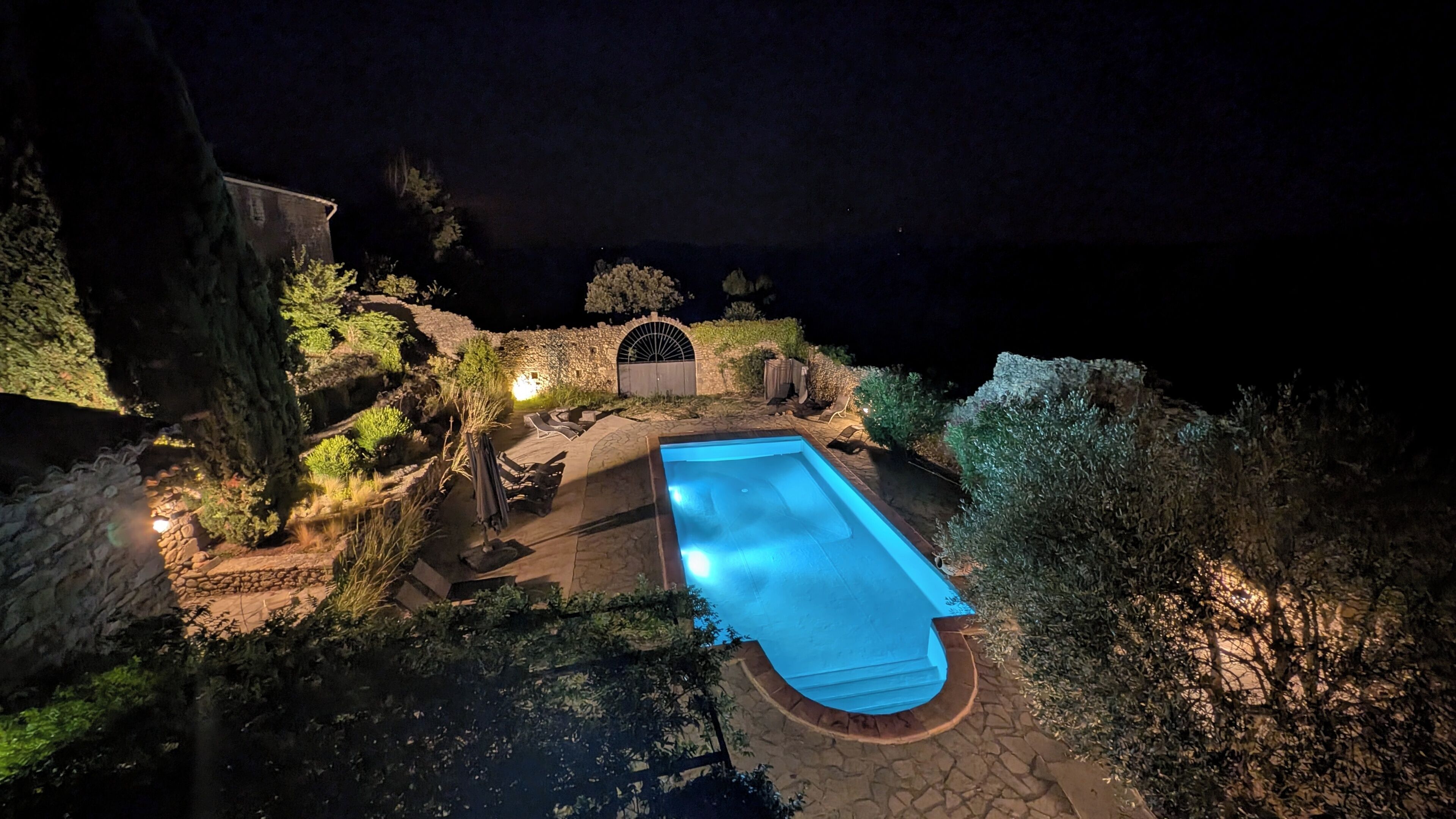 Vista nocturna de una piscina rodeada de muros de piedra y vegetación, con un arco de piedra y un área de estar al aire libre.