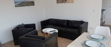 A modern living room with a black sofa, a smaller black armchair, a white coffee table, and a dining table set for two with white plates and glasses.