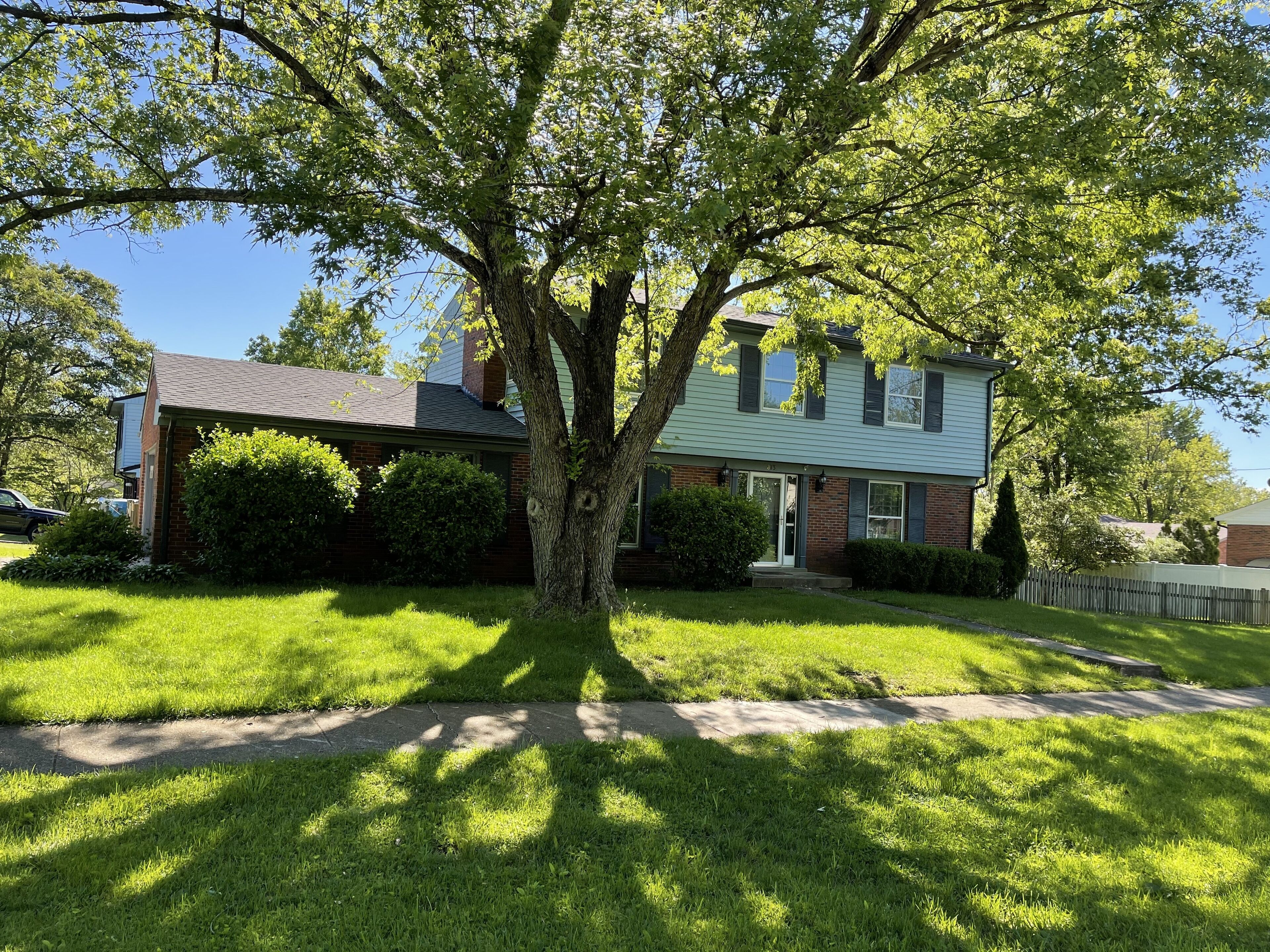 A two-story house with a large tree in front, a well-maintained lawn, and a wooden fence.