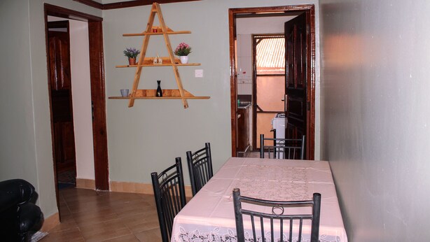 Zona pranzo con tavolo imbandito per quattro persone, scaffale a scaletta in legno con piante in vaso e vista su un'altra stanza attraverso una porta aperta.