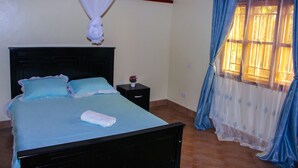 Un letto rifatto con cura, con copriletto e cuscini azzurri, un comodino con una piccola pianta e una finestra con tende blu.