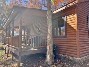 A wooden cabin with a porch, a window with shutters, and a tree in front.