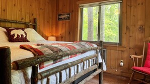 Un dormitorio de cabaña de madera con una cama, un sillón mecedora, una ventana con vista a los árboles y un cartel en la pared.