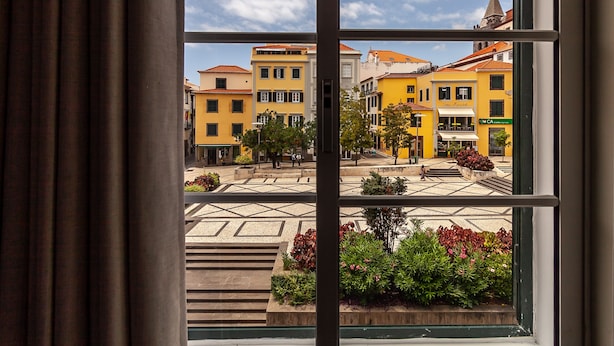 A view through a window of a courtyard with buildings, a café, and a church. - Zarco B&B - Bed and Breakfast (Funchal)
