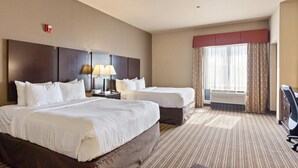 A hotel room with two beds, a desk, a lamp, a chair, and a large window with curtains. - Portland Inn & Suites (Portland)