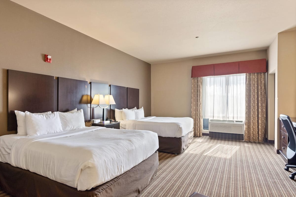 A hotel room with two beds, a desk, a lamp, a chair, and a large window with curtains.