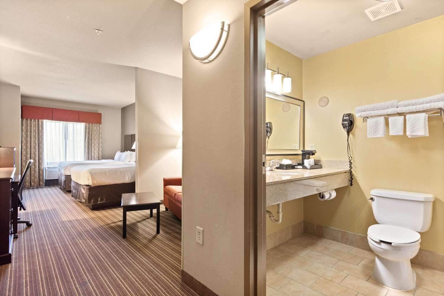 A hotel room with a bed, a desk, a chair, a small table, a bathroom with a sink, a mirror, a toilet, and a towel rack.
