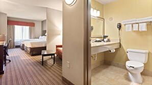 A hotel room with a bed, a desk, a chair, a small table, a bathroom with a sink, a mirror, a toilet, and a towel rack. - Portland Inn & Suites (Portland)