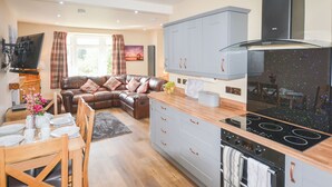 A modern kitchen with a built-in stove, oven, and grey cabinets. There is a dining area with a wooden table and chairs, and a brown leather sofa in the background.
