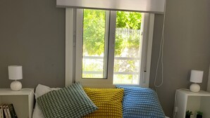 En seng med hvidt sengetøj, tre puder i grøn, gul og blå, samt et vindue med en hvid rullegardin.