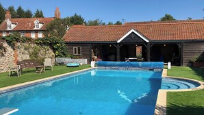 Una piscina rectangular con borde de baldosas, rodeada por un césped y un edificio de madera con techo de tejas.