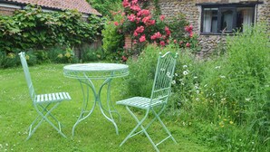 Un jardín con una mesa metálica redonda y dos sillas, rodeado de abundante follaje y plantas en flor.