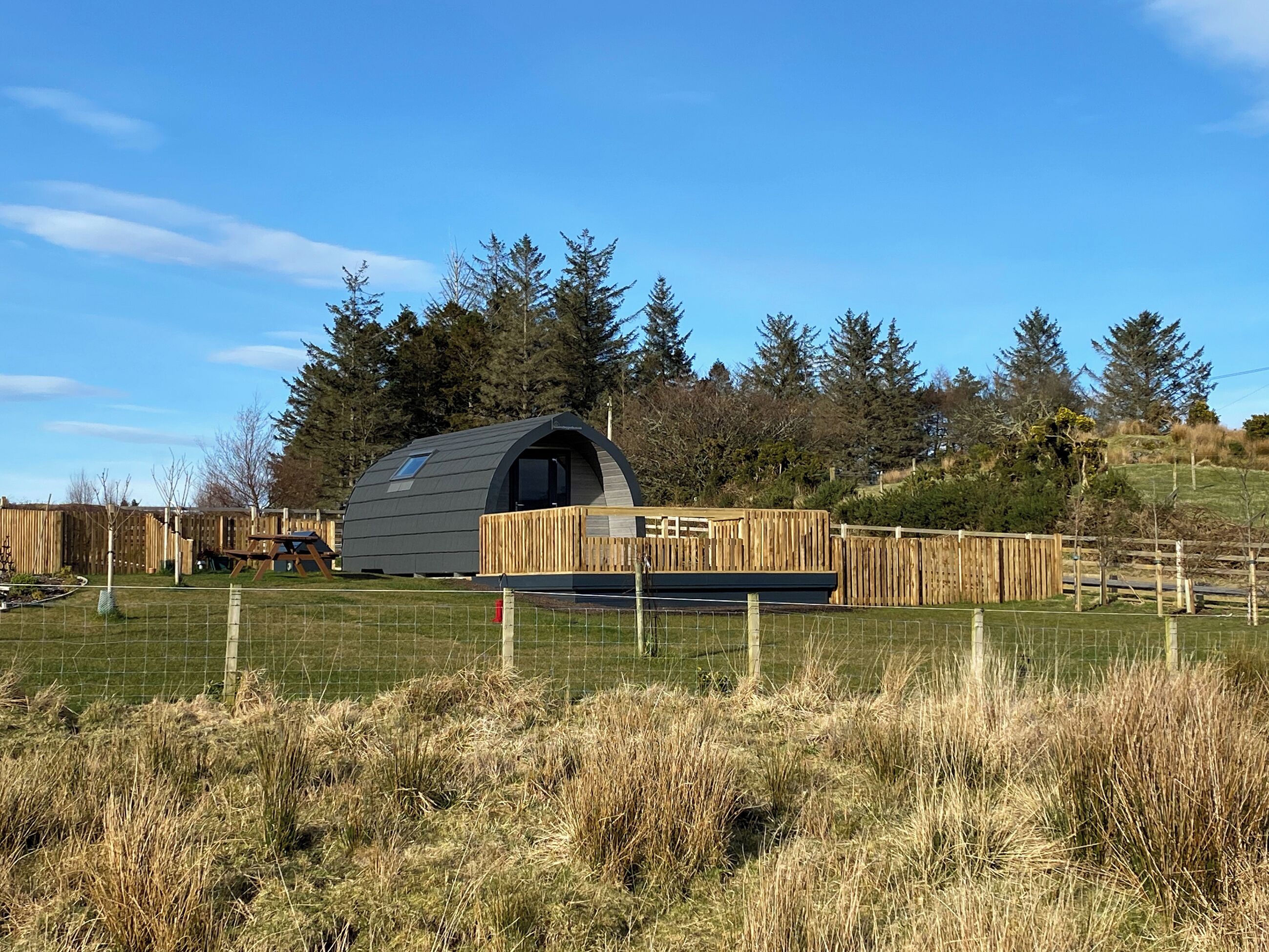 A small, semi-circular wooden structure with a sloped roof, surrounded by a wooden fence and a grassy field.