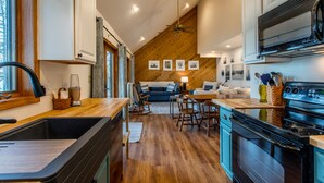 A compact kitchen with a stove, sink, and wooden flooring. There is a dining area with a table and chairs, and a sofa in the background.