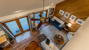 A spacious living area with a ceiling fan, wooden flooring, a sofa, a coffee table, and a view through large windows.
