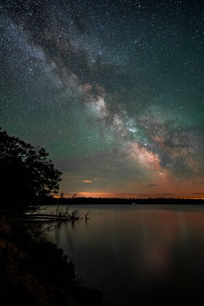 A night sky filled with stars and the Milky Way over a calm body of water with a silhouette of trees.