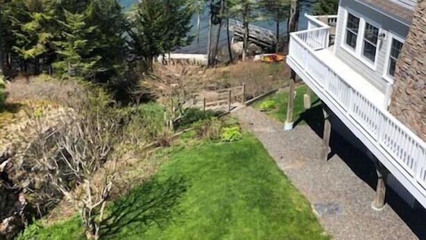 A house with a stone foundation, a white exterior, and a balcony overlooking a lake surrounded by trees.