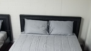 Una cama con estructura negra y ropa de cama blanca, una pared de ladrillo blanco y suelo de madera.