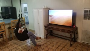 Una sala de estar con televisor sobre un mueble de madera, una silla colgante y una cocina al fondo.