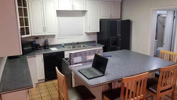 Una cocina con armarios blancos, un refrigerador negro, un microondas y una laptop sobre la encimera.